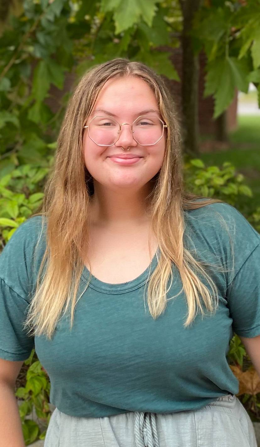 farren outside in a teal shirt and gold-rimmed glasses smiling infront of a short brick wall and a green tree