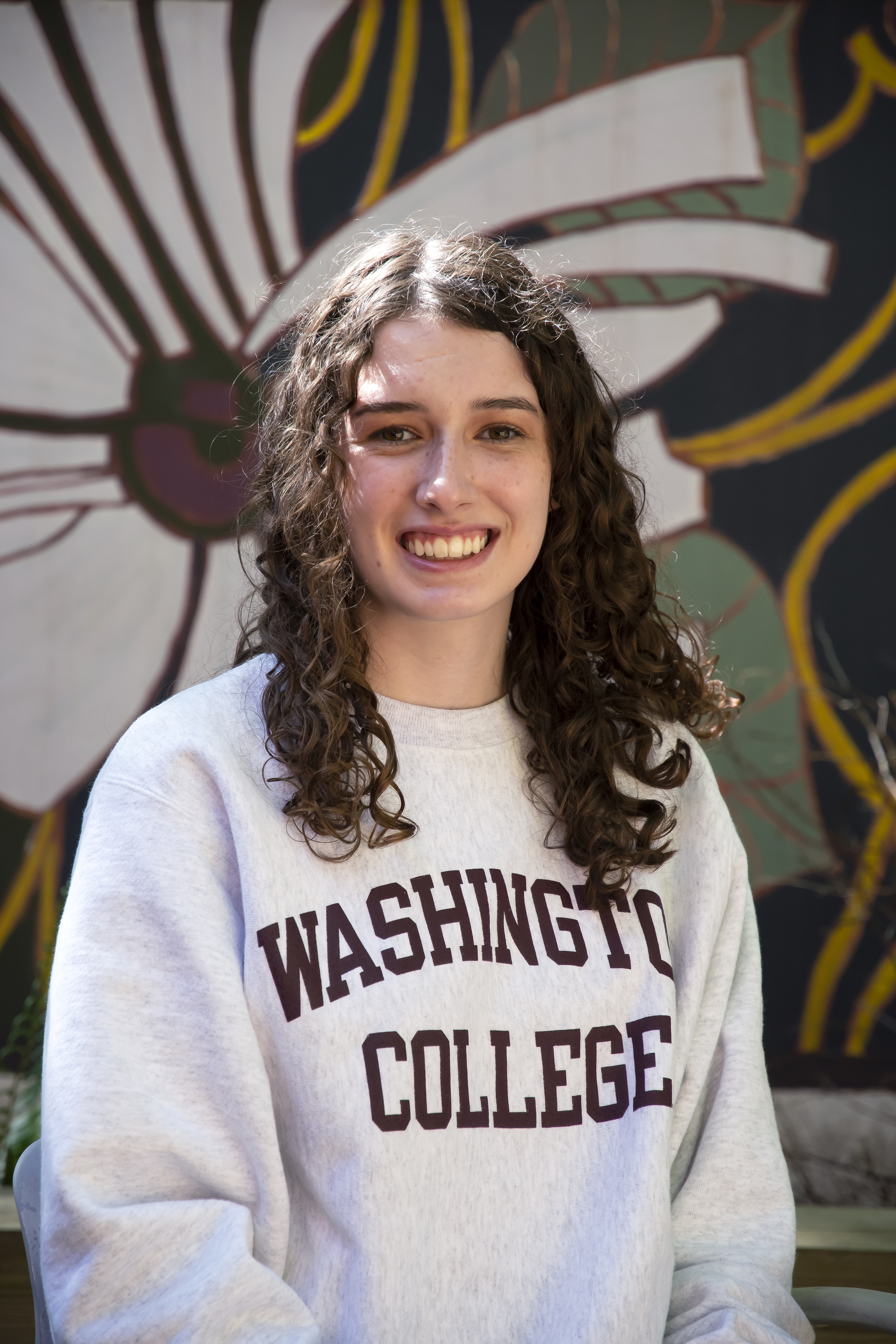 Emma, smiling and wearing a white Washington College sweatshirt with a painted mural behind her. 