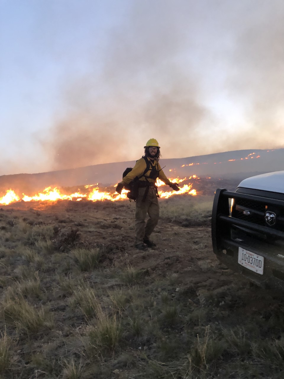 Lanning, wearing protective fire gear, smiling in a field with fire behind him. 